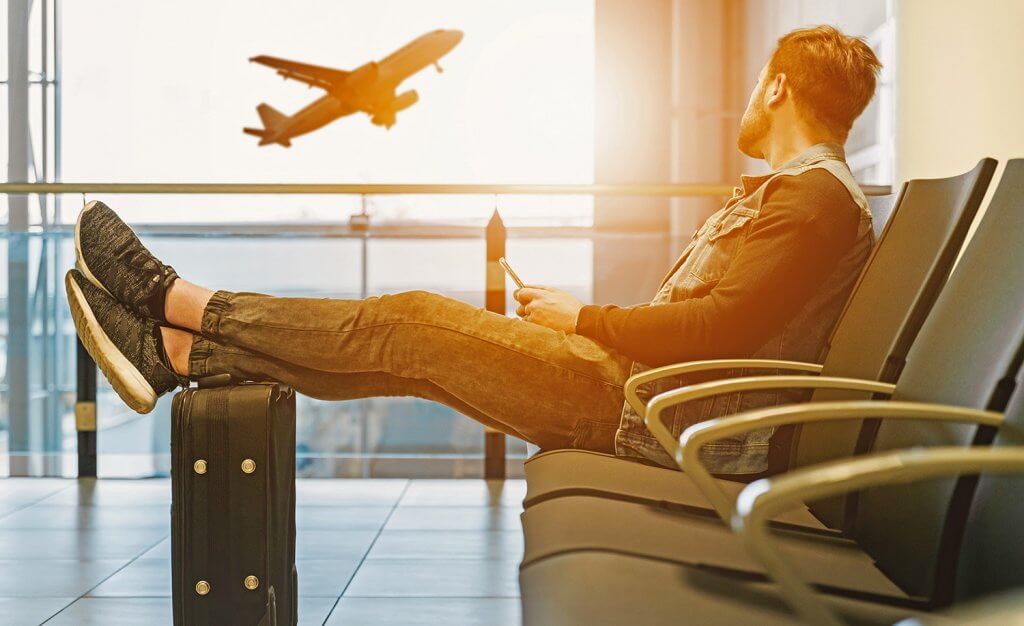 Traveler sitting in airport with feet on luggage set looking at an airplane